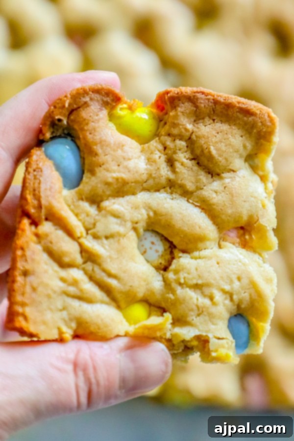 A hand reaching for a slice of gooey Easter blondie, generously topped with Cadbury Mini Eggs and M&Ms, illustrating its rich and inviting texture.