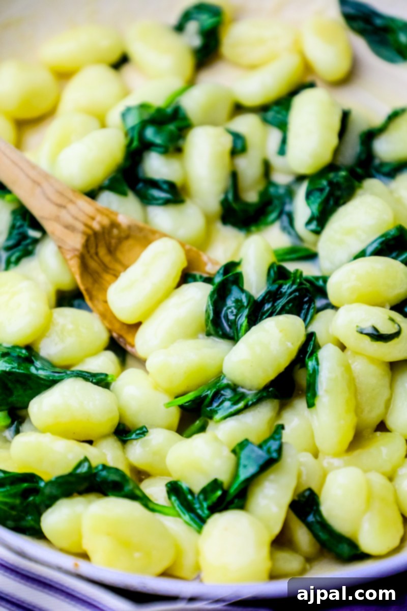A wooden spoon stirs tender gnocchi and wilted spinach in a creamy Parmesan sauce within a single pan, illustrating the ease of this one-pot recipe.
