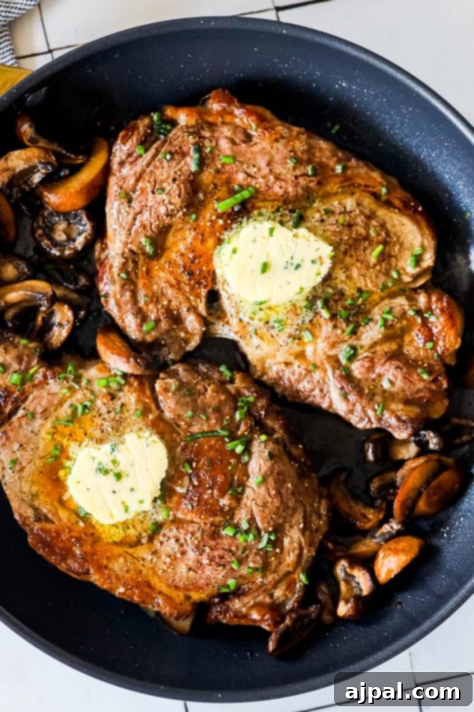 Succulent ribeye steaks in a skillet with sautéed mushrooms and melting butter pats, fresh from the oven broiler.