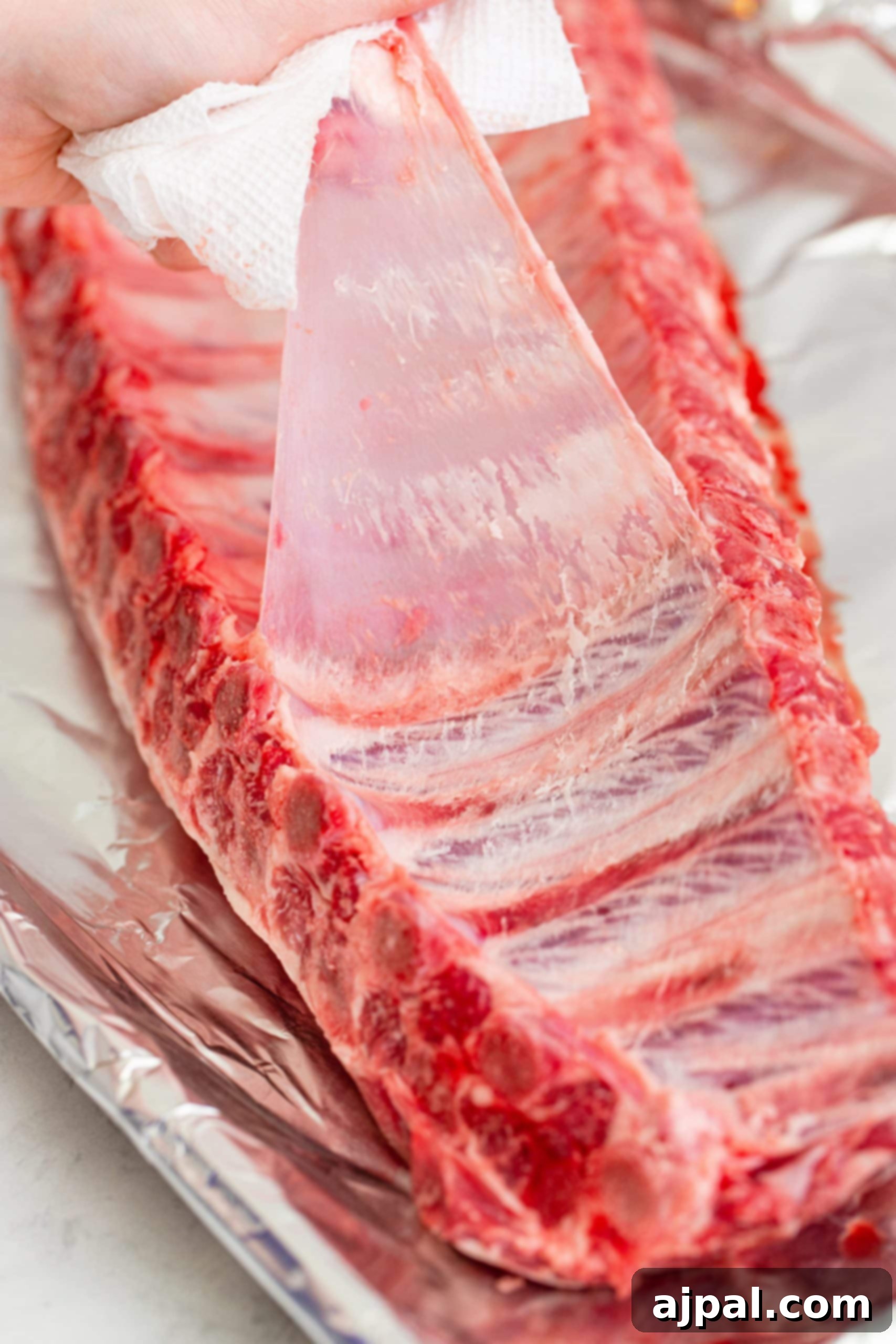 A hand expertly removing the silver skin membrane from a rack of raw pork ribs, a crucial step for tender results.