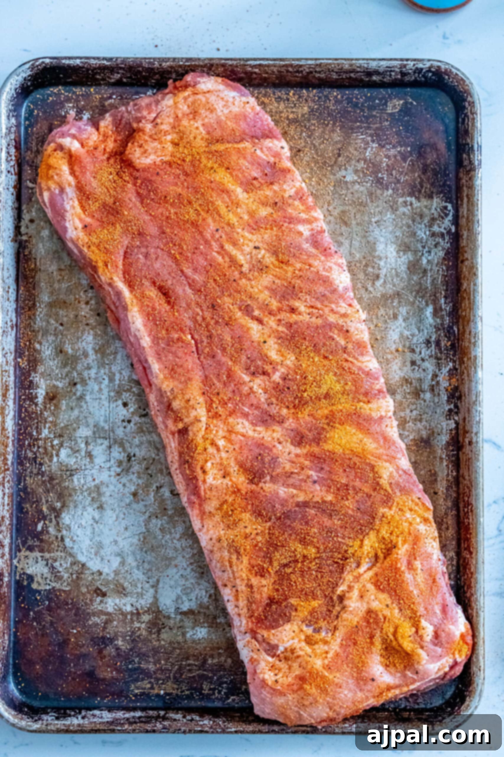 A baking sheet holds a rack of pork ribs generously coated in a rich, savory dry rub, ready for the smoker.