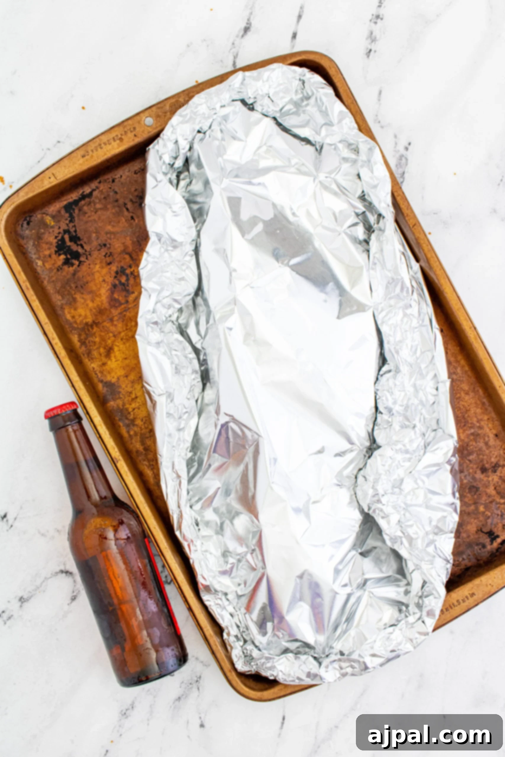Pork ribs tightly wrapped in aluminum foil on a baking sheet, with a bottle of beer nearby, ready for the next stage of cooking.