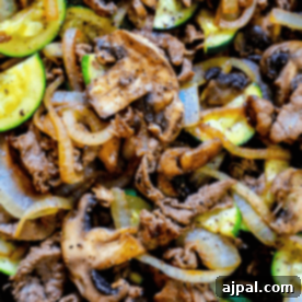 Close-up of a cooked dish featuring slices of beef, mushrooms, onions, and zucchini, seasoned with black pepper. This easy sirloin skillet with vegetables recipe is both savory and satisfying.