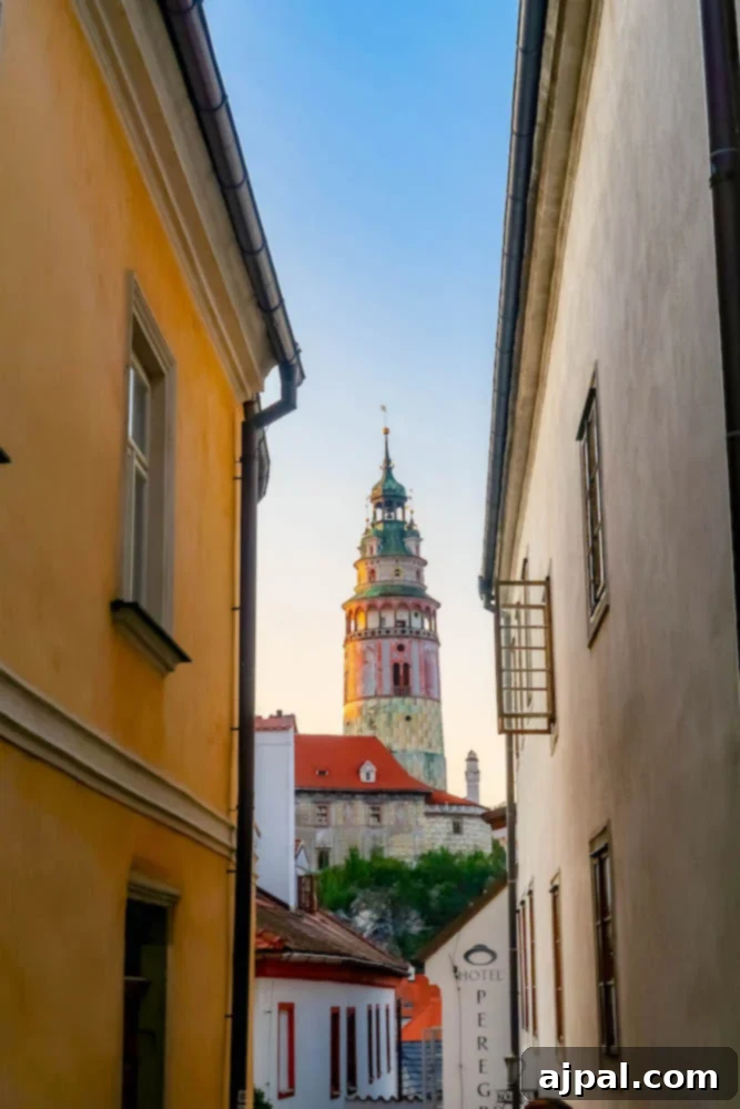 Narrow cobblestone alleyway in Cesky Krumlov with historic buildings and the iconic castle tower visible in the background, hinting at the town's medieval heritage.