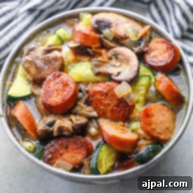 A bowl of keto sausage and vegetable soup recipe, featuring sliced sausage, mushrooms, zucchini, carrots, onions, and savory broth, sits on a gray surface with a striped cloth in the background.