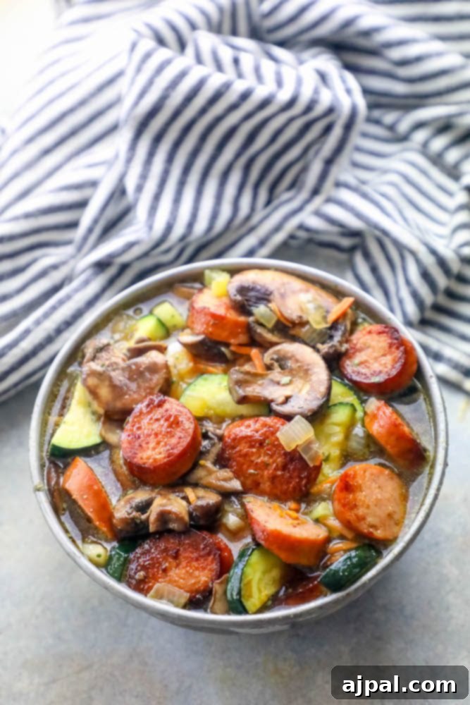 An appetizing overhead shot of Andouille sausage, various mushrooms, and thinly sliced zucchini bathing in a rich, savory broth within a deep soup bowl, highlighting the fresh ingredients.