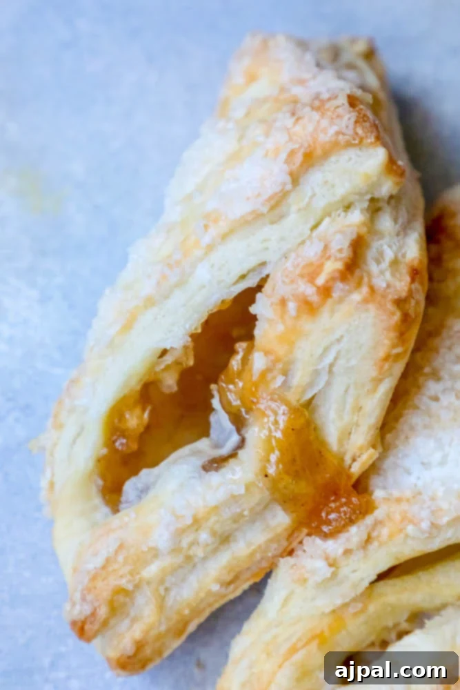 Two easy puff pastry apple turnovers, adorned with a delicate dusting of powdered sugar and a side of apricot jam, presented on a rustic wooden table.