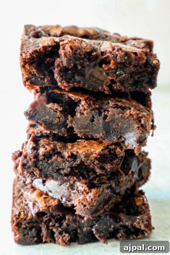 Five thick, glossy fudge brownies stacked invitingly on a white surface, showcasing their irresistibly gooey interiors and a drizzle of hot fudge sauce.
