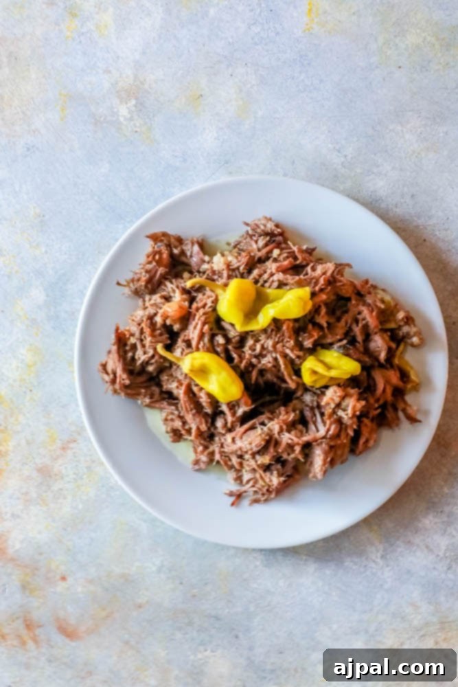 A perfectly plated serving of Slow Cooker Mississippi Roast with bright yellow pepperoncini peppers on a white plate on a kitchen counter.