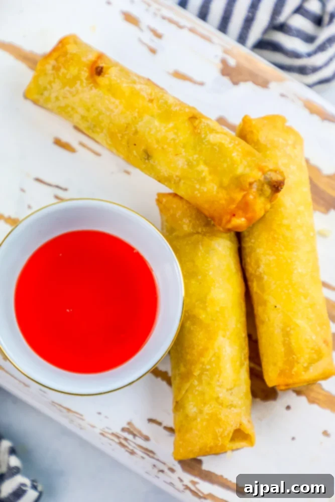 Crispy Homemade Egg Rolls 5 Three golden-brown egg rolls arranged neatly next to a small bowl of classic sweet and sour dipping sauce.