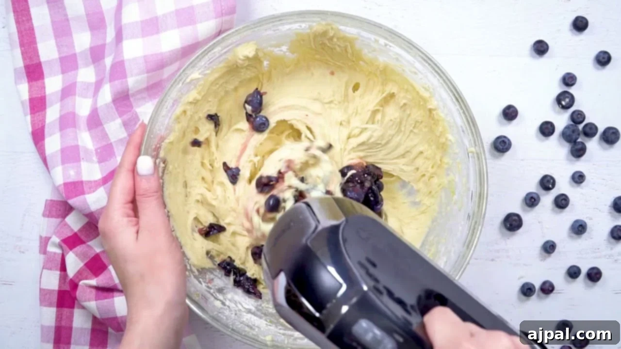 A person using a hand mixer to mix blueberries in a bowl for The Best Blueberry Muffins recipe.