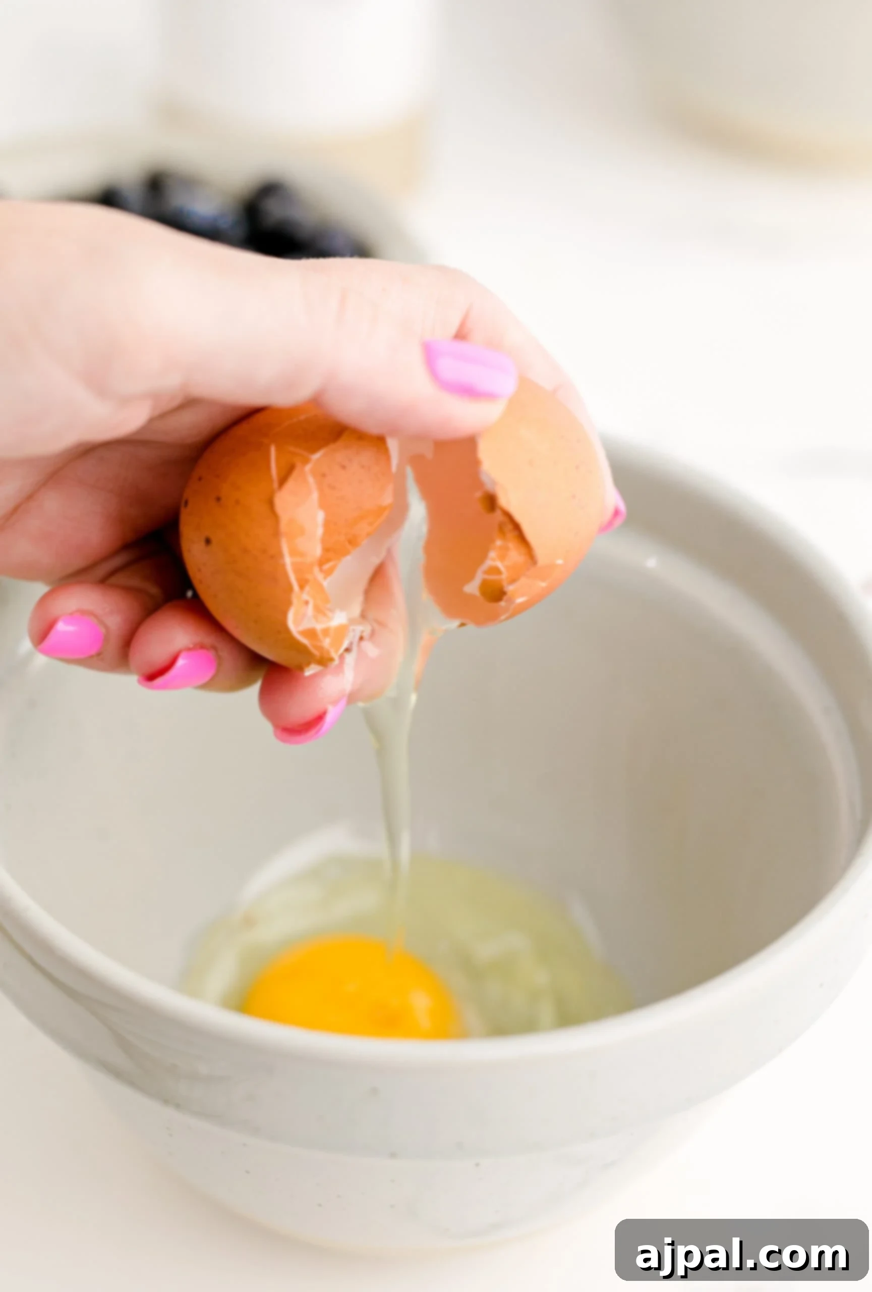 A person preparing a jumbo blueberry muffin recipe by breaking an egg into a bowl.