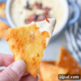 A hand holding a triangular cheese cracker dipped in a creamy sauce evokes the essence of keto nachos, with more crispy treats and indulgent dips in the background.