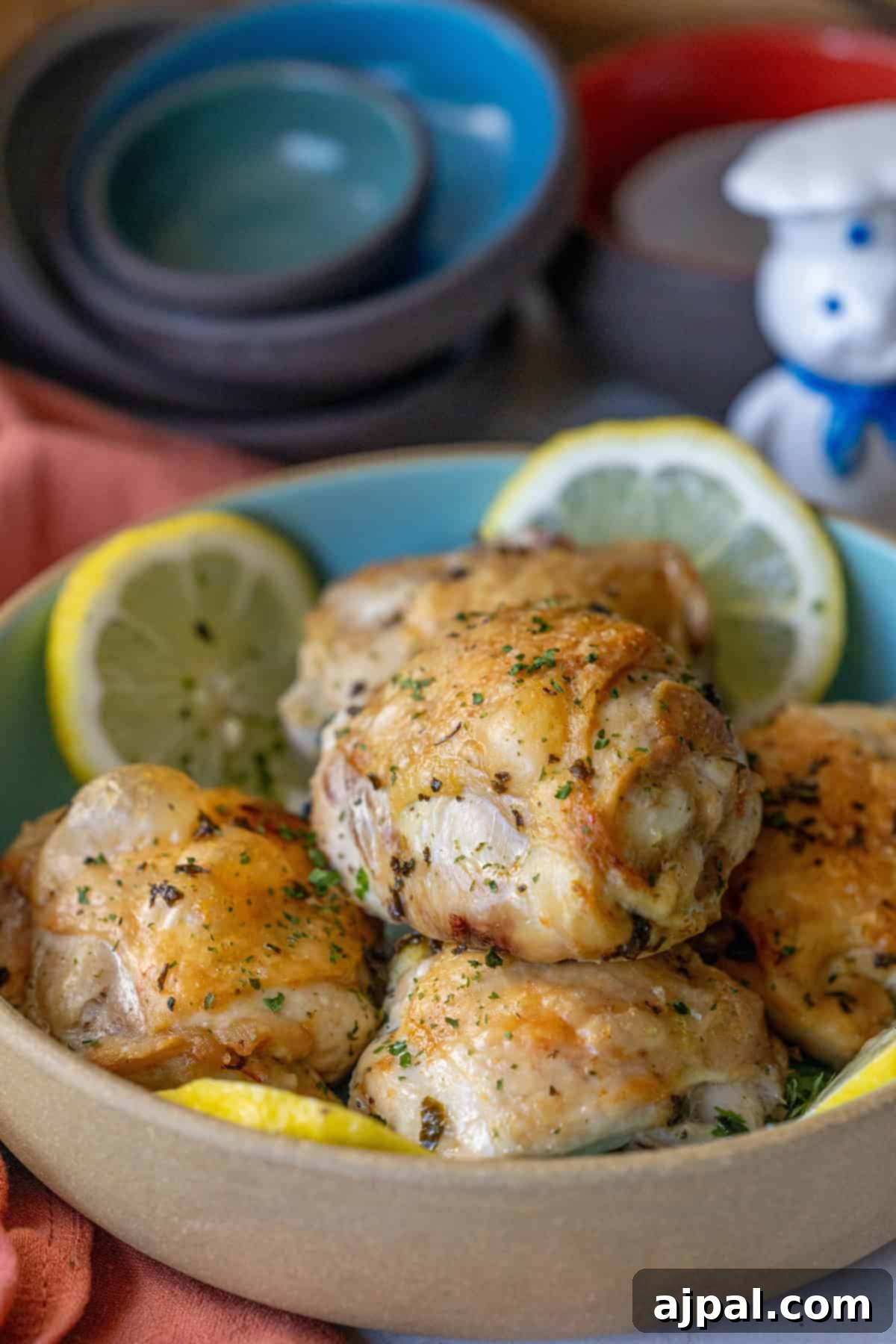 A bowl of seasoned air fried lemon chicken thighs garnished with parsley and lemon slices, with stacked bowls and a figurine in the background.