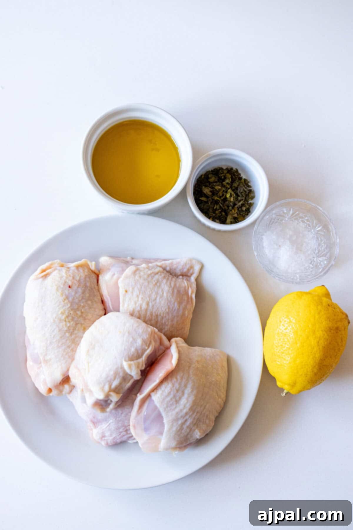 Raw chicken thighs rest on a plate alongside a lemon, a bowl of olive oil, a bowl of herbs, and salt on a white surface—perfect for crafting an air-fried lemon chicken bursting with flavor.