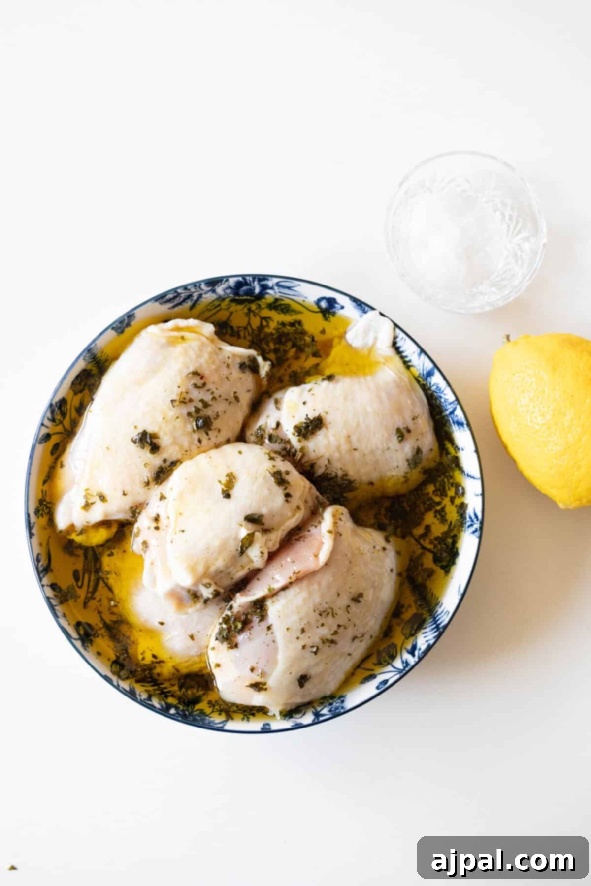 Raw chicken marinating in herbs and oil in a blue and white bowl, ready to become a delicious air fried lemon chicken. Nearby, a whole lemon and a small bowl of salt await their turn in this easy air fryer lemon chicken recipe.