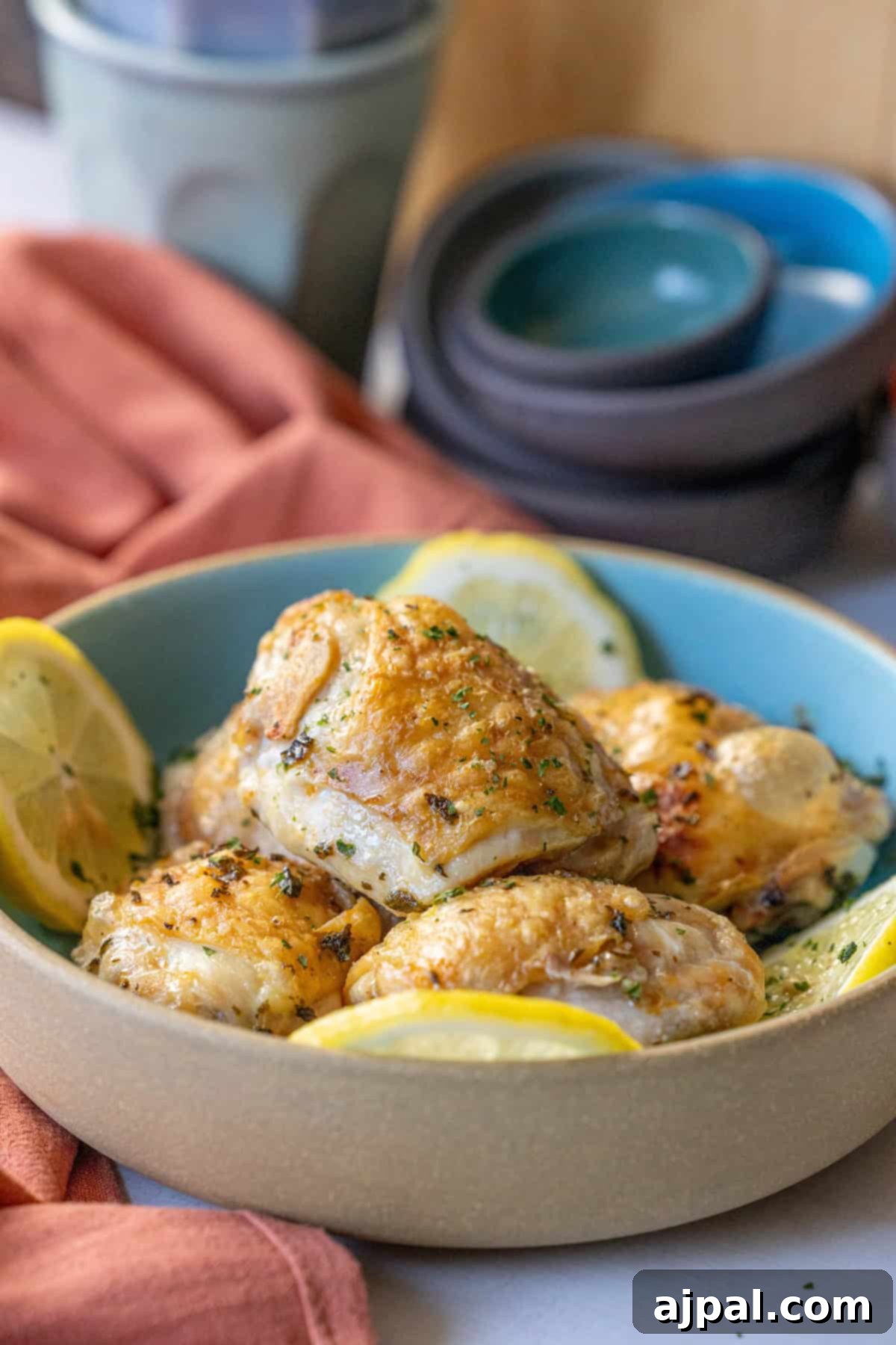 A bowl of easy air fryer lemon chicken thighs, garnished with lemon slices and herbs, sits enticingly in the foreground. Several stacked bowls and a cup linger in the background, setting the scene for this flavorful dish.
