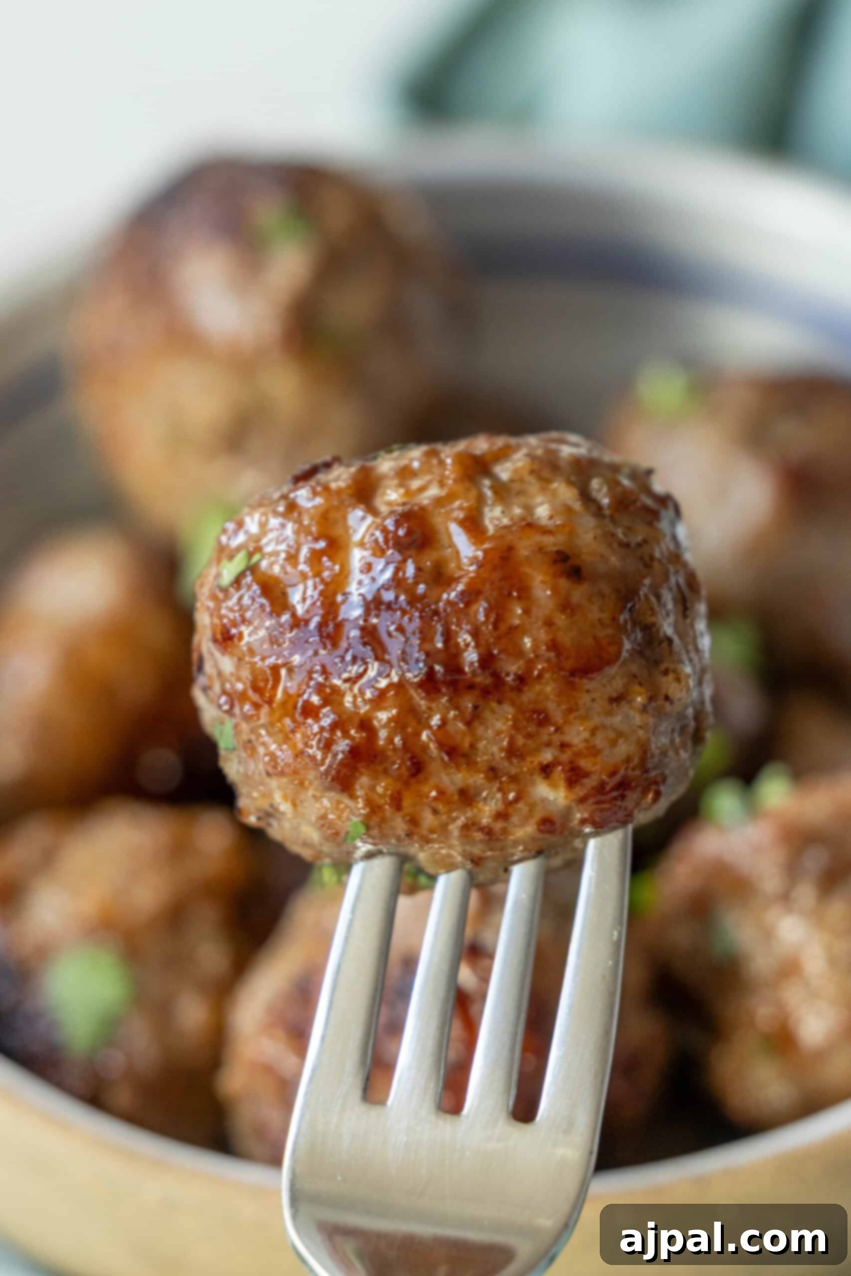 Close-up of a fork holding a perfectly browned, glazed air fried meatball against a backdrop of more meatballs in a bowl.