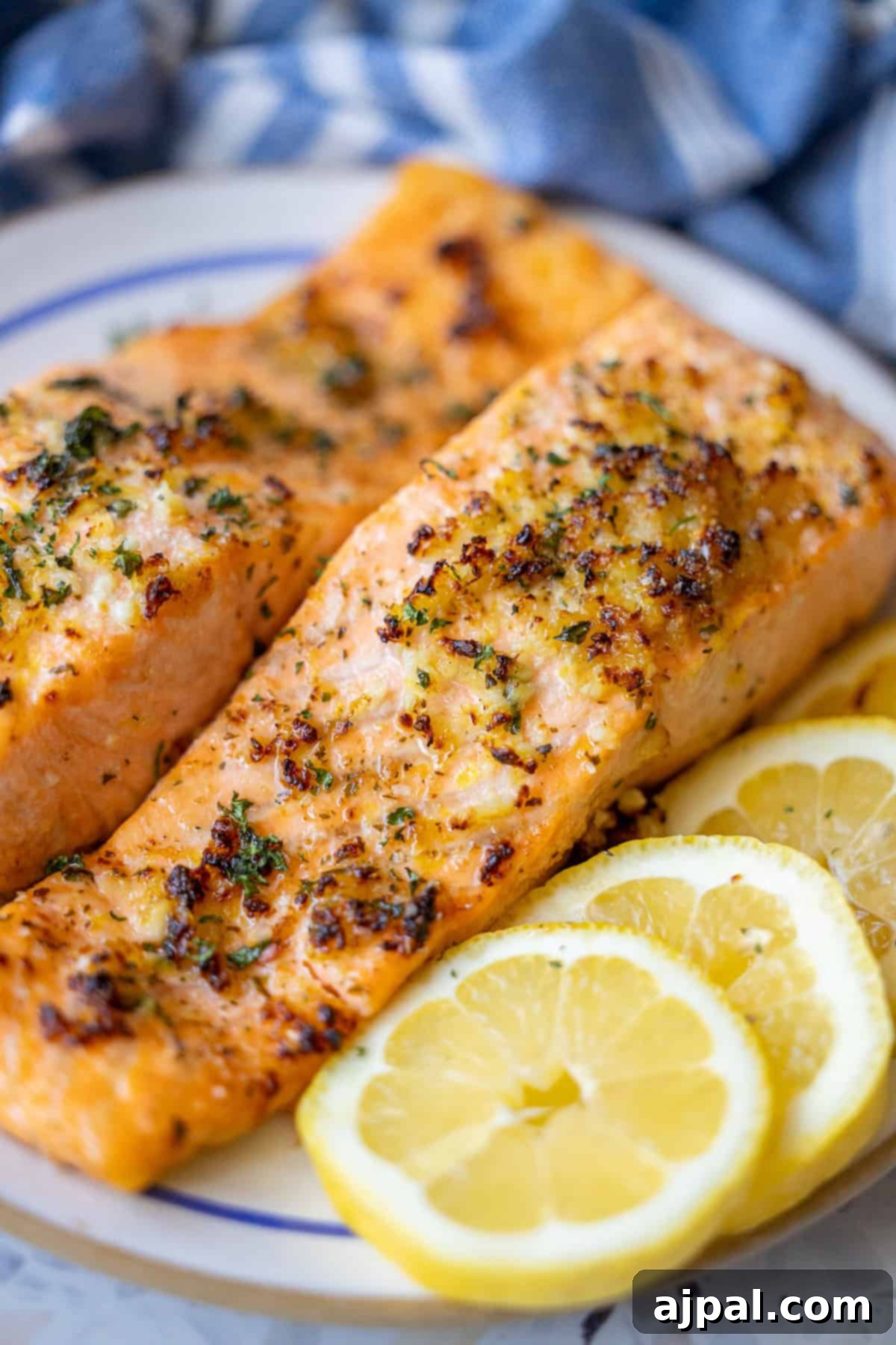 Easy Air Fryer Lemon Garlic Salmon 5 Two cooked air fryer salmon fillets with herbs are served on a plate next to several slices of fresh lemon. A blue and white cloth is in the background, highlighting this Easy Air Fryer Lemon Garlic Salmon Recipe.