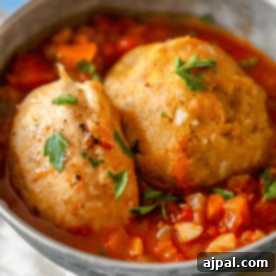 Two matzo balls in a tomato-based broth with diced vegetables, garnished with parsley, served in a gray bowl with chicken mofongo.