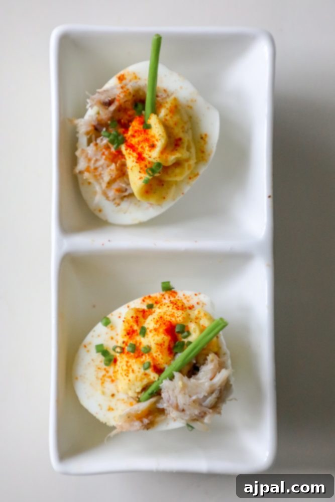 Spicy Crab Bloody Mary Deviled Eggs 3 A pair of beautifully arranged Bloody Mary deviled eggs, each topped with delicate crab meat and a vibrant sprinkle of herbs, presented on a pristine white plate.