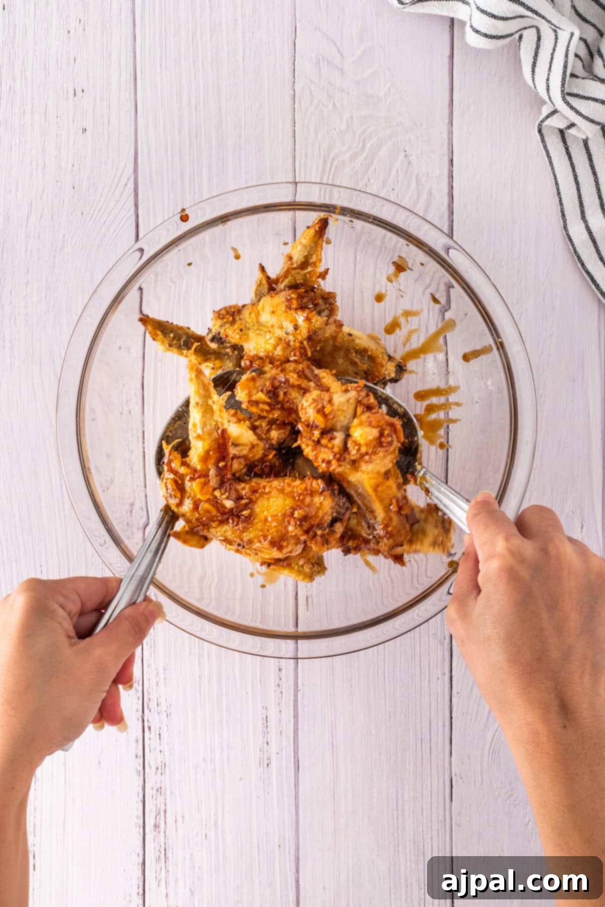 Crispy Oven Wings 8 Two hands mix honey-glazed chicken wings in a glass bowl with metal tongs on a white wooden surface.