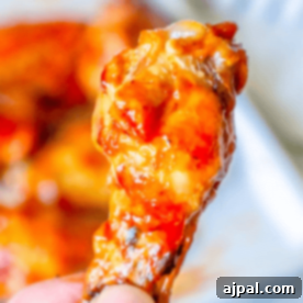Close-up of a hand holding a chicken wing coated in a thick, reddish-orange sauce from our Baked Crispy BBQ Chicken Wings recipe, with more sauced wings blurred in the background.
