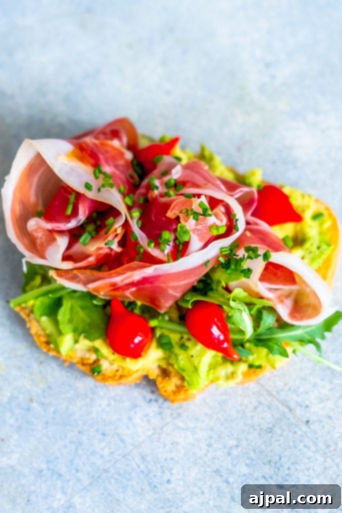 A delectable piece of keto bread topped with smashed avocado, thin slices of prosciutto, fresh arugula, chopped peppers, and chives, inviting a healthy and flavorful breakfast.