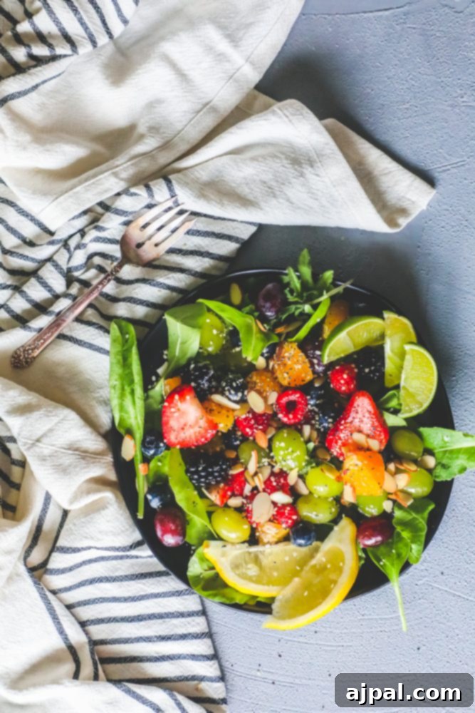 A fresh spinach and fruit salad with a variety of colorful fruits like grapes, oranges, strawberries, adorned with almonds and poppyseeds, presented in a glass bowl.