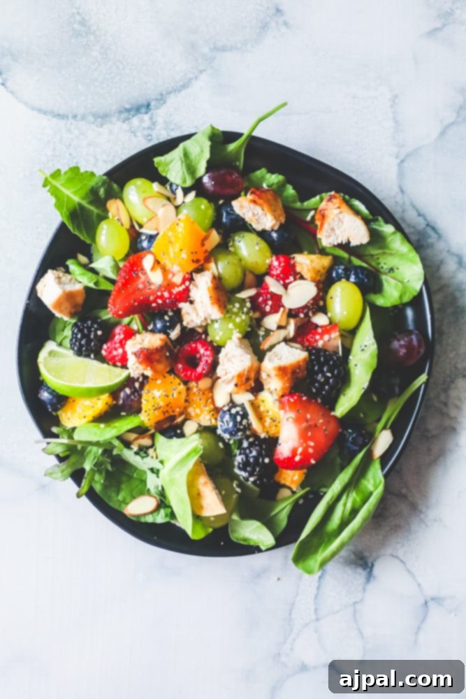 Vibrant spinach salad featuring grapes, strawberries, blackberries, blueberries, raspberries, tender chicken, poppy seeds, hemp hearts, and a glistening citrus poppyseed dressing on a black plate, ready to be enjoyed.