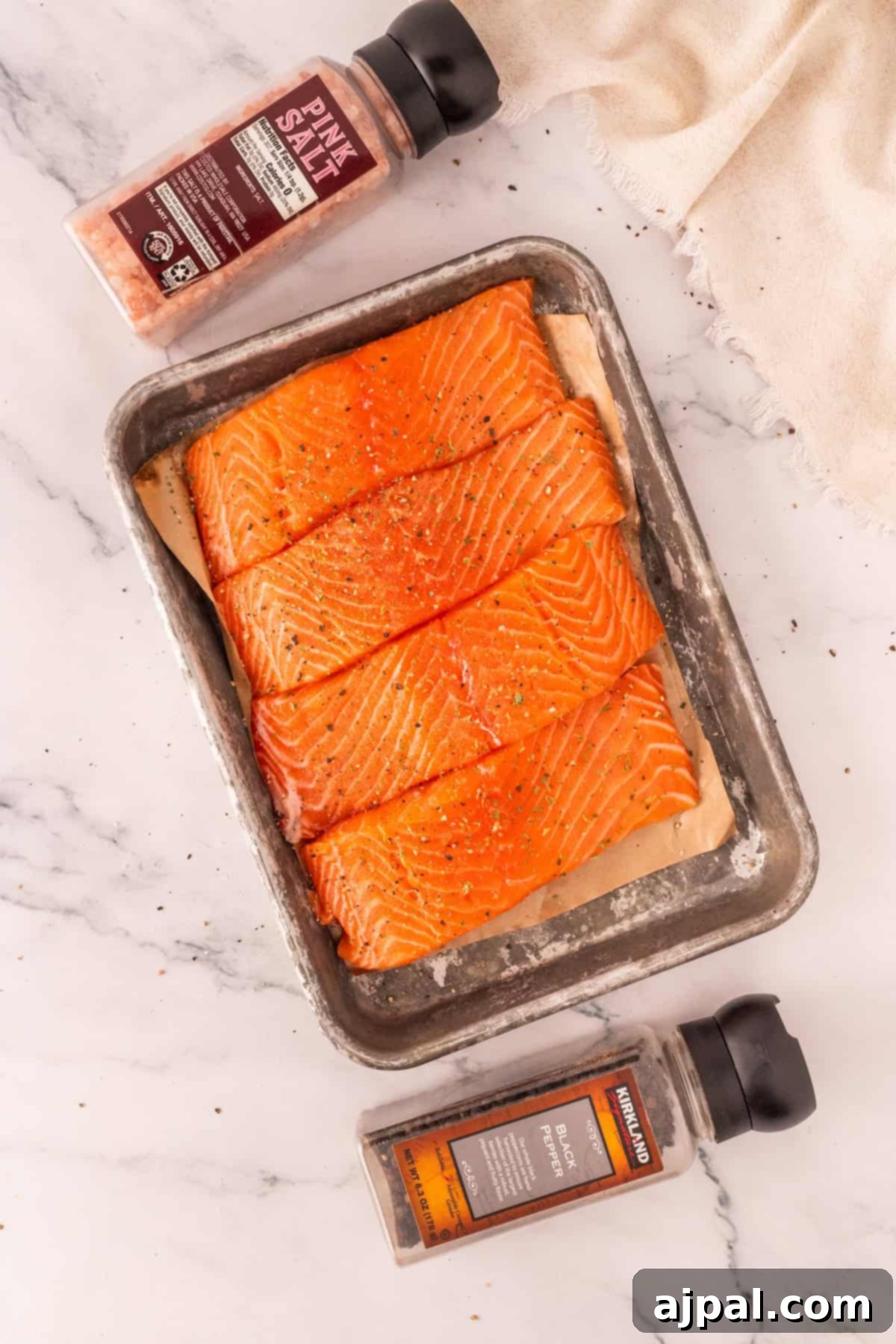 Effortless Oven-Baked Salmon 3 Raw salmon fillets on a metal tray, sprinkled with spices, ideal for the Best Baked Salmon. Bottles of pink salt and black pepper are placed nearby on a marble surface, perfect for trying a garlic baked salmon recipe.