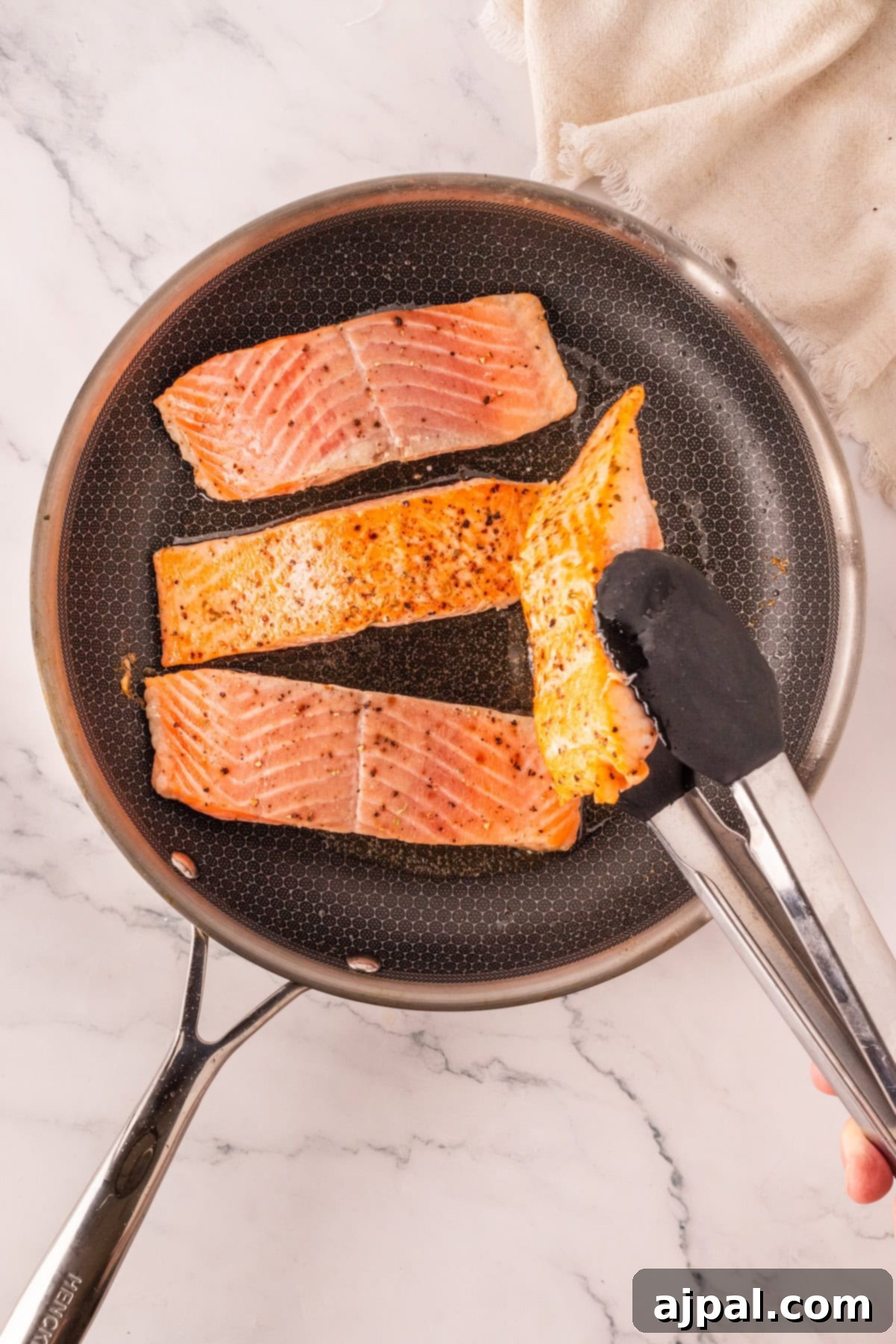 Effortless Oven-Baked Salmon 4 Three seasoned salmon fillets are being cooked in a nonstick skillet, with one fillet being lifted by metal tongs—perfect for anyone seeking the best baked salmon recipe with a delicious garlic twist.