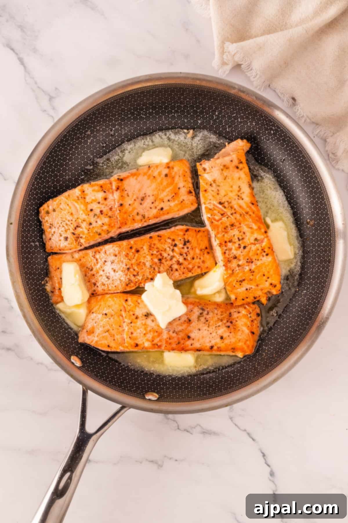 Effortless Oven-Baked Salmon 5 Four seared salmon fillets topped with melting butter are cooking in a nonstick skillet on a marble countertop—perfect inspiration for the best baked salmon recipe you'll want to try at home.