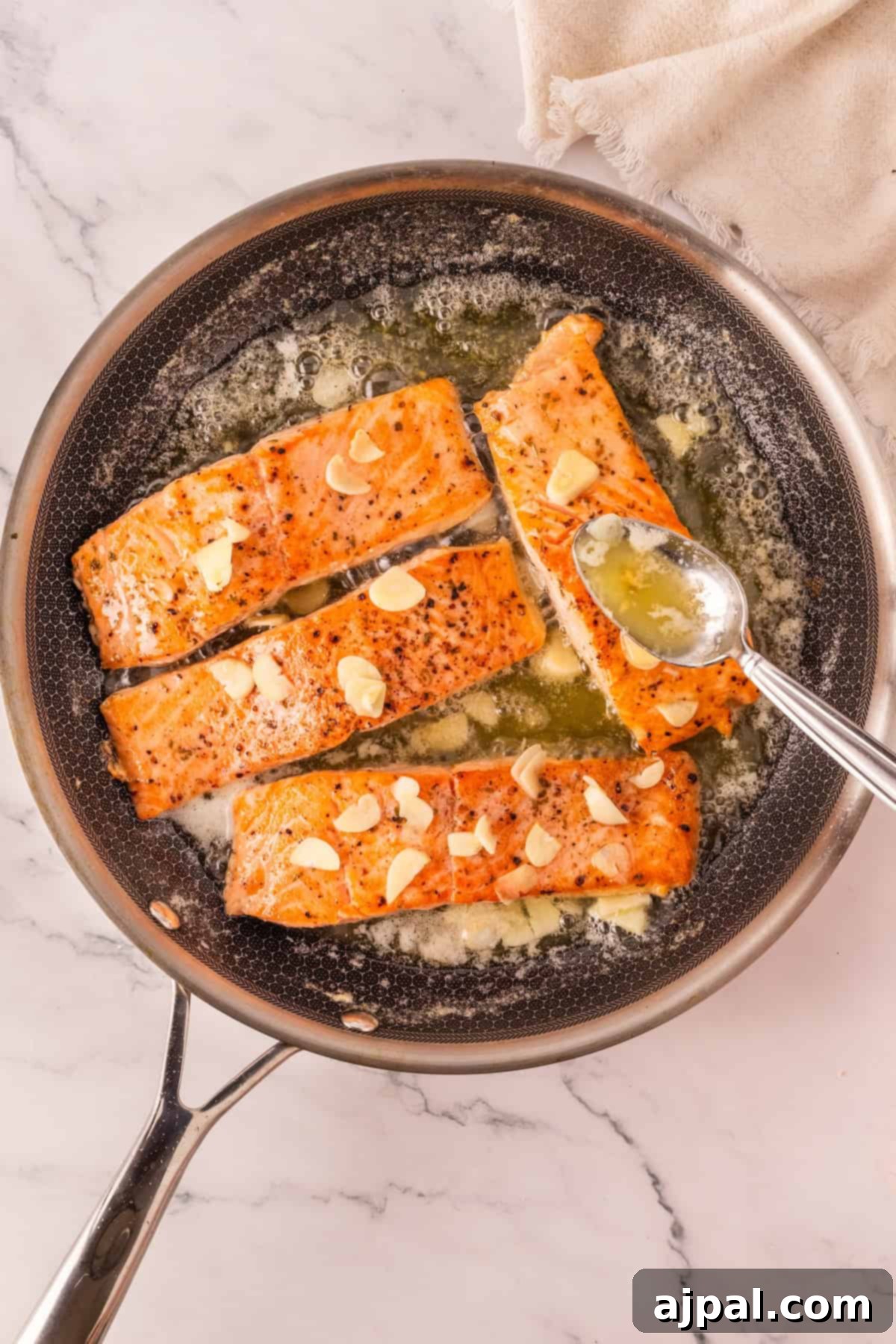 Effortless Oven-Baked Salmon 6 Four seared salmon fillets topped with sliced garlic are cooking in a skillet with melted butter, as a spoon drizzles butter over the fish for an irresistible garlic baked salmon recipe.