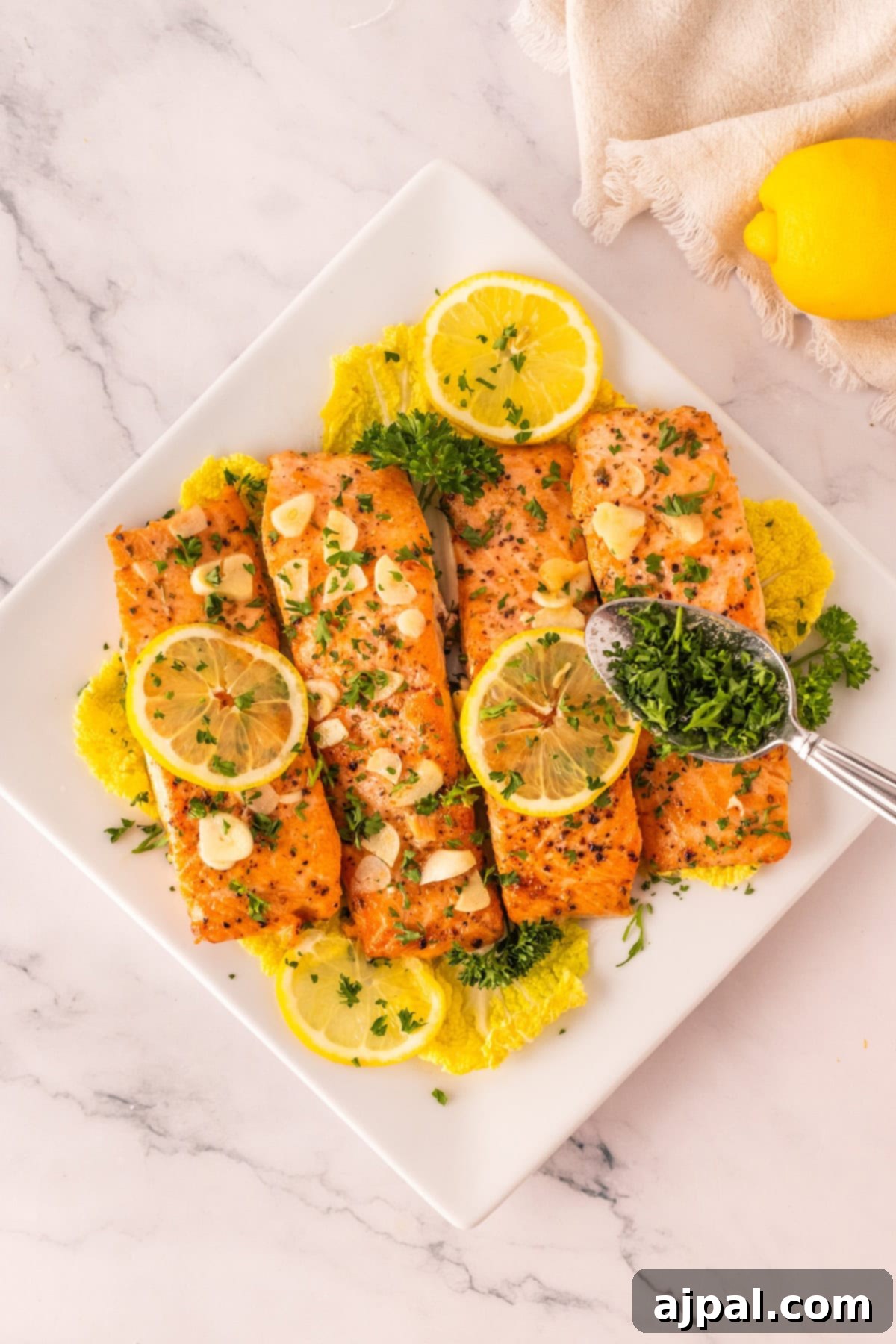 Effortless Oven-Baked Salmon 7 Four baked salmon fillets topped with lemon slices and chopped herbs are arranged on a white plate with leafy greens, as a spoon adds more herbs—a tempting glimpse of the best baked salmon recipe. A lemon and napkin sit nearby.