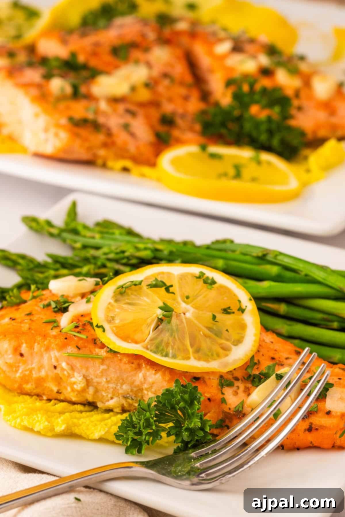 Effortless Oven-Baked Salmon 9 A plate of cooked salmon garnished with lemon slices and parsley, served with asparagus and a fork, showcases the best baked salmon recipe, with another plate of salmon featured in the background.