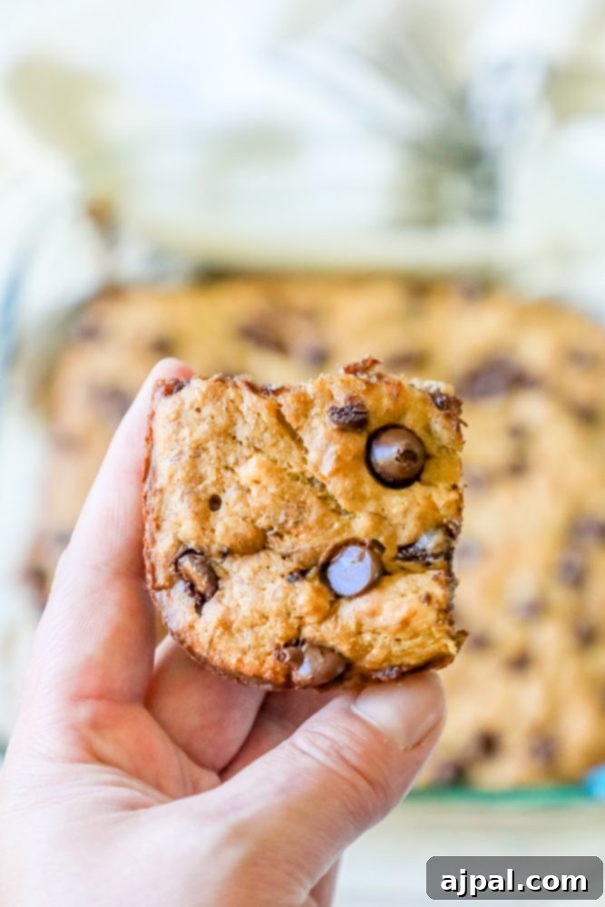 A close-up shot of a single banana chocolate chip bar, held up to reveal its moist texture and abundant chocolate chunks, with a full pan of bars blurred in the background.