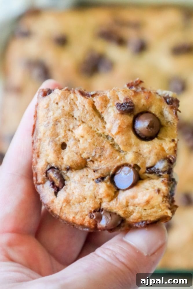 A person's hand holding a single, generously chocolate-studded banana bar, showcasing its inviting golden color and soft appearance.
