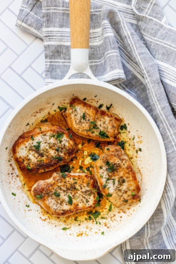 Perfectly Pan Fried Thin Pork Chops 3 A close-up view of two glistening, pan-fried pork chops cooking in a hot skillet, surrounded by aromatic herbs.