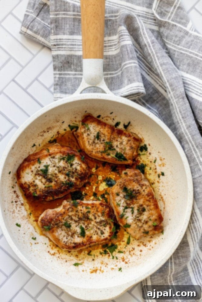 Perfectly Pan Fried Thin Pork Chops 9 A view of two pan-fried pork chops, perfectly seared and golden, arranged neatly in a dark skillet.