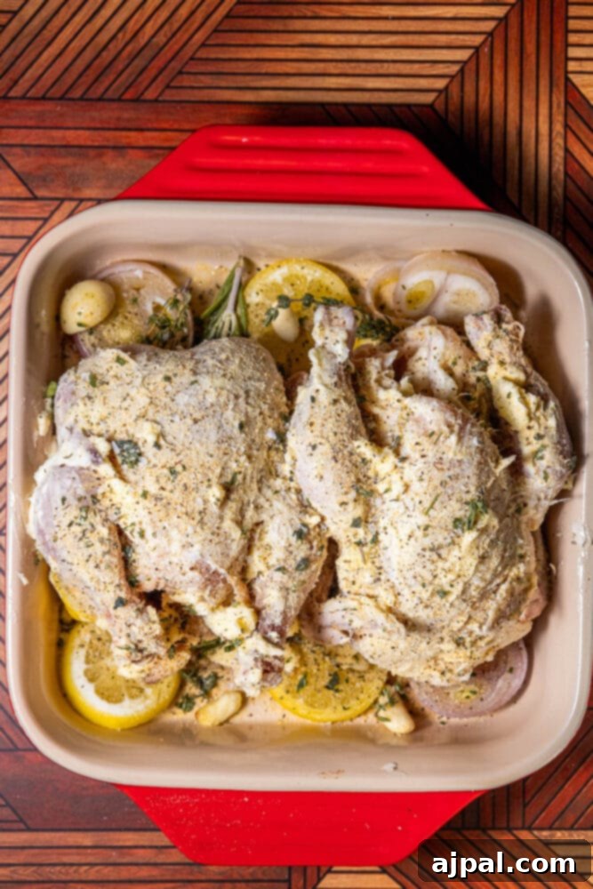 Raw Cornish game hens, covered in butter and herbs, arranged on lemon, shallot, and herbs in a baking dish.