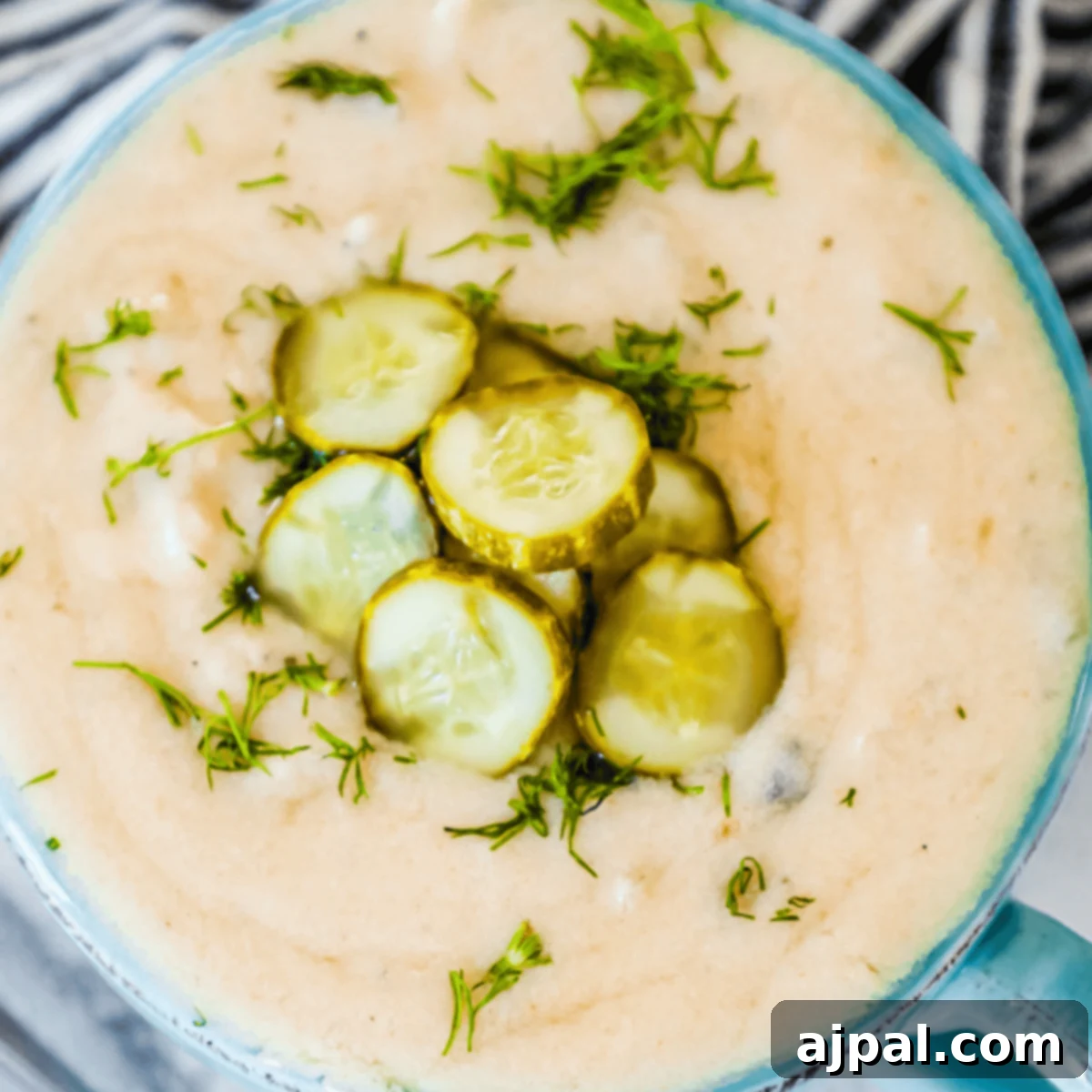 Simple Dill Pickle Soup 4 A close-up of a creamy pickle soup garnished with sliced pickles and chopped fresh herbs in a blue bowl—a delicious twist on the classic dill pickle soup recipe.