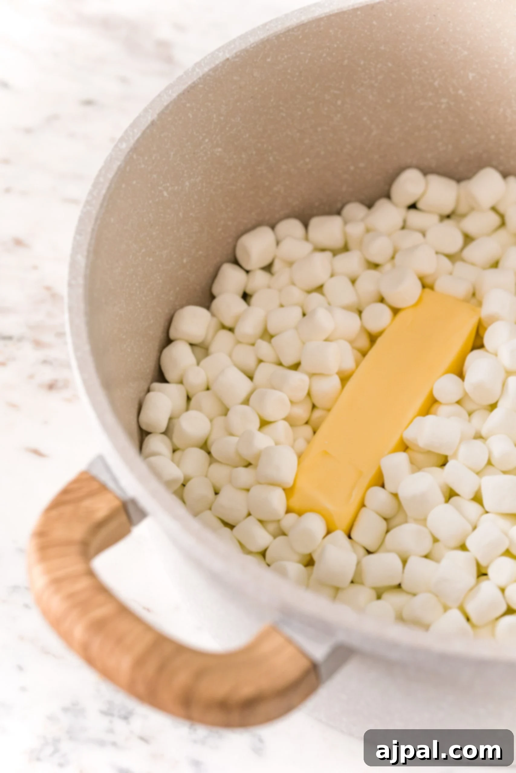 A saucepan on a stovetop, containing cubes of butter and a generous amount of mini marshmallows, ready to be melted together.