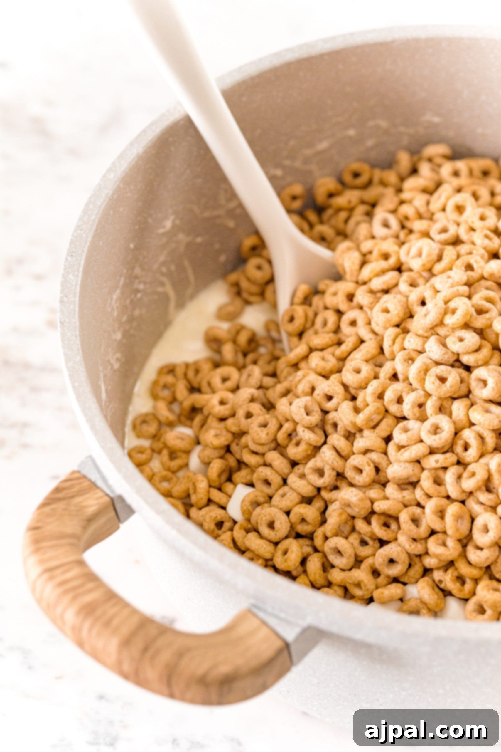 A large quantity of Honey Nut Cheerios being stirred into a pot of melted butter and marshmallow mixture, ensuring every cereal piece is thoroughly coated.