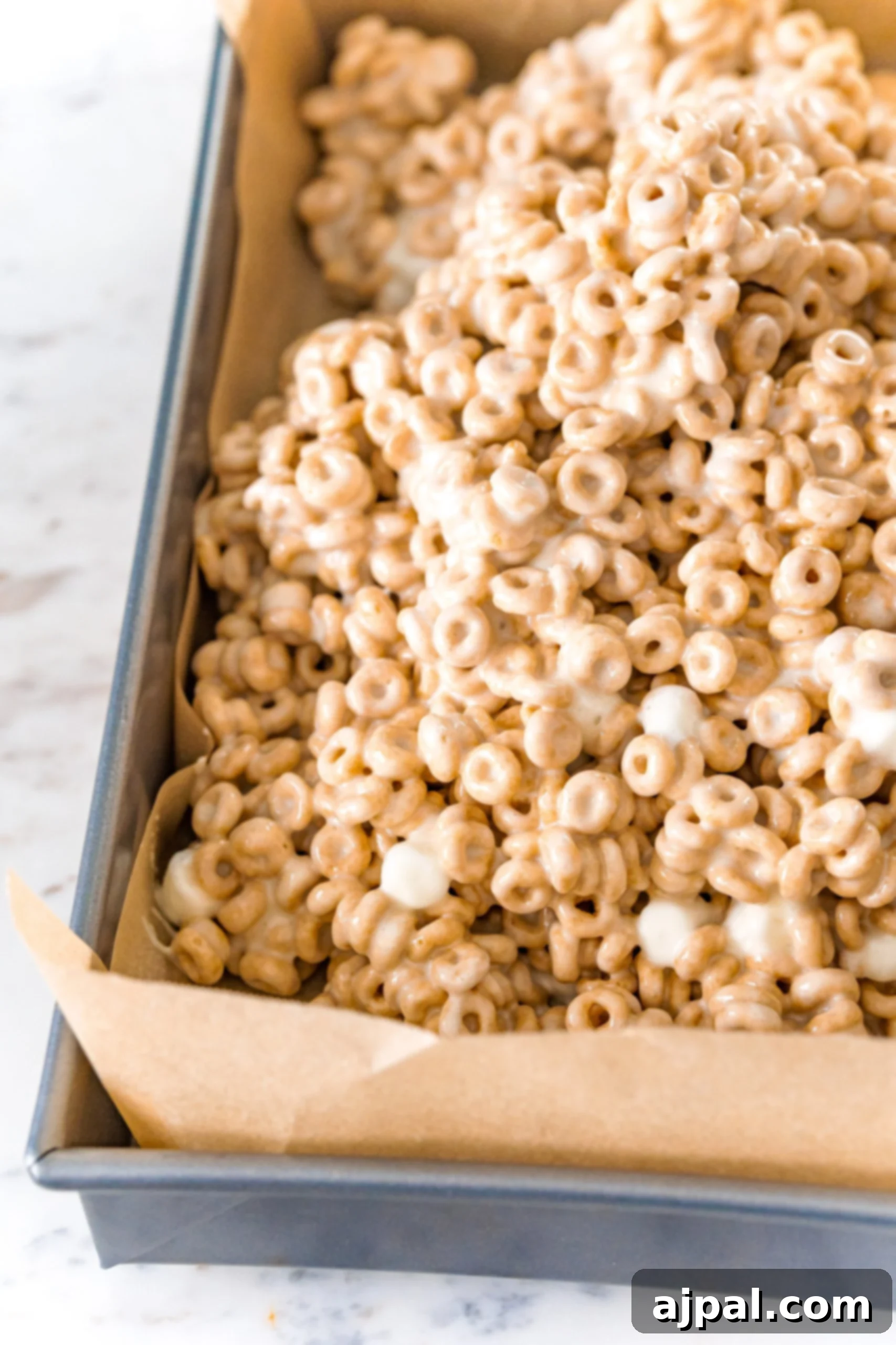 A close-up of the Cheerio mixture being gently pressed into a parchment-lined 9x13 inch baking pan, ready for setting.