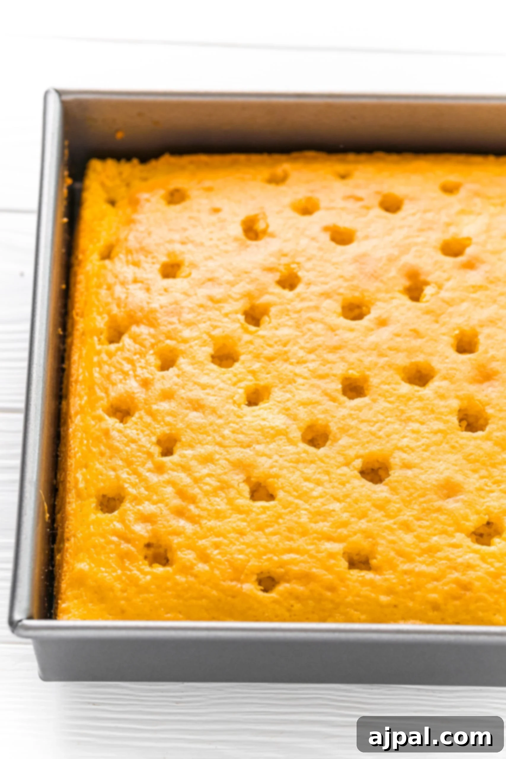 A close-up shot of the baked lemon cake, still in its pan, with a wooden spoon handle being used to poke evenly spaced holes across its warm surface.