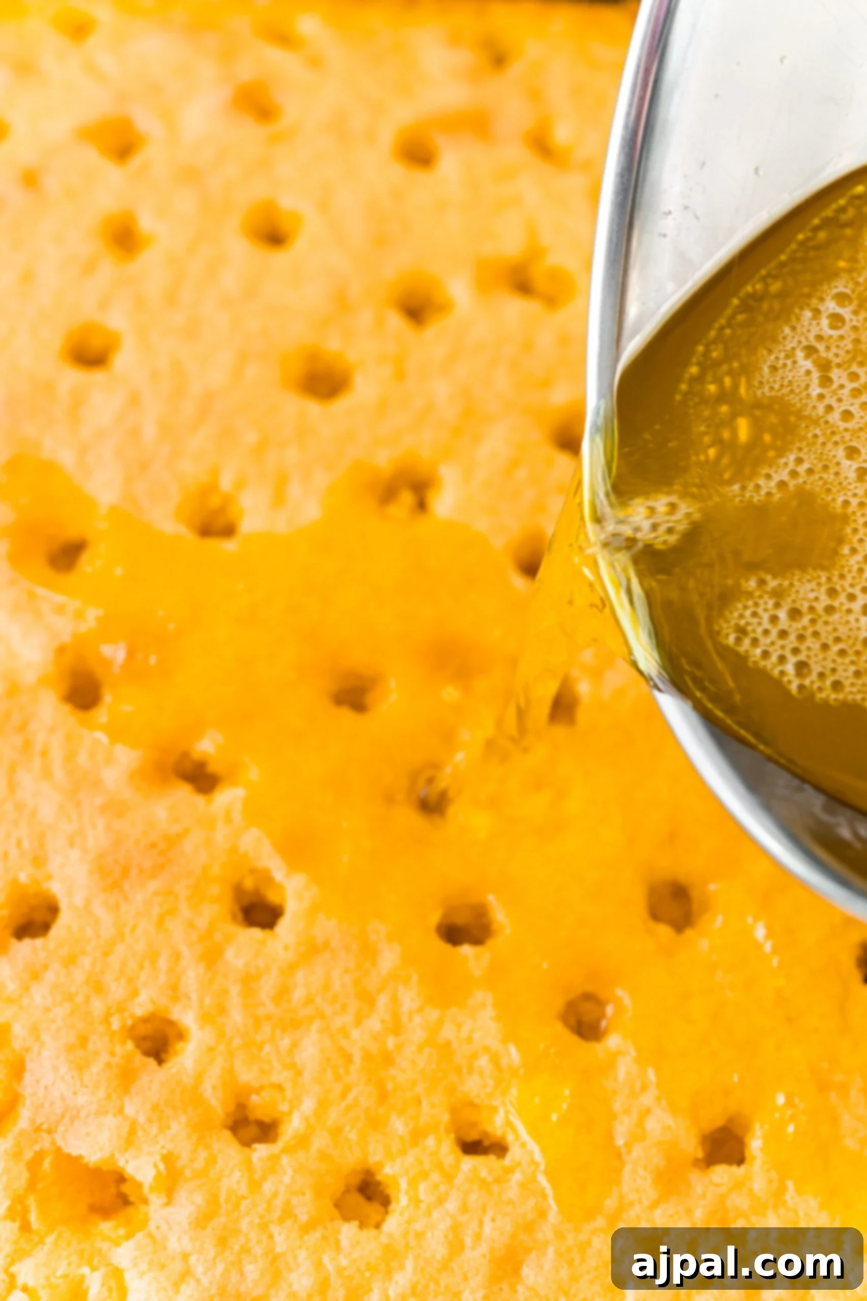 The vibrant yellow lemon Jello mixture is being carefully poured over the top of a freshly poked lemon poke cake, filling each hole with zesty goodness.