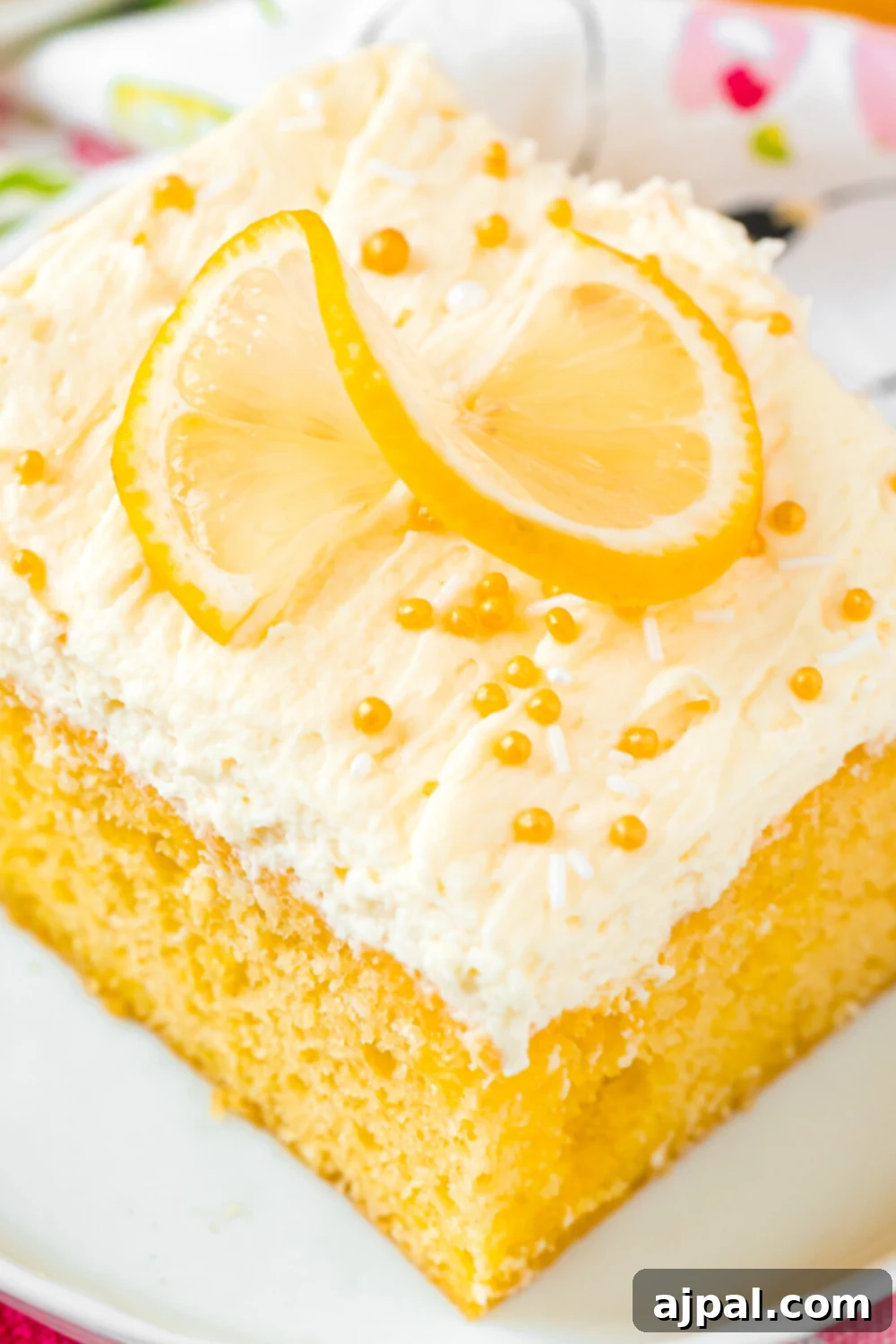 A beautifully frosted slice of Lemon Jello Poke Cake, garnished with fresh lemon zest and a hint of lemon slice, presented on a white plate, ready to be enjoyed.