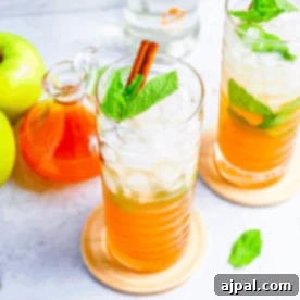 Two tall glasses of iced orange drink with mint leaves and cinnamon sticks, next to green apples, a glass jar of orange liquid, and a bottle—perfect inspiration for an Apple Cider Mojitos Cocktail recipe.