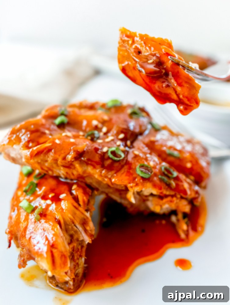 Instant Pot Sticky Asian BBQ Ribs 5 A fork is gently lifting a piece of tender, saucy Asian BBQ rib from a white plate, showcasing its succulent texture.